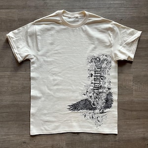 Puede incluir: Una camiseta de manga corta color crema con un estampado gráfico negro en la parte inferior derecha. El diseño muestra una pila vertical de elementos ornamentales en espiral, un par de alas detalladas en la base y texto caligráfico intrincado.