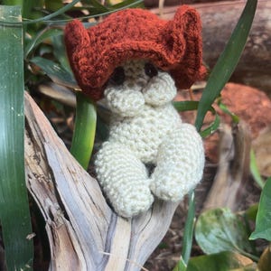 May include: A small, handmade crochet plush figure shaped like a mushroom. The cap is a deep, textured rust-orange color with a ruffled edge, while the stem and base are a creamy off-white color. The figure features two small, dark, round eyes.