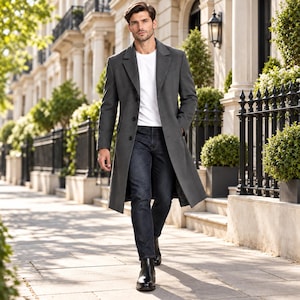 May include: A charcoal gray long overcoat worn over a plain white t-shirt and dark denim jeans. The coat features a notched lapel, a three-button front closure, and long sleeves. The outfit is completed with black leather ankle boots.