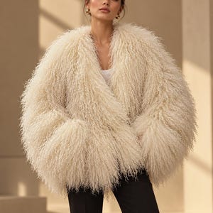 May include: A cream-colored, long-haired, curly-textured jacket with a deep V-neckline and long, voluminous sleeves. The jacket is worn over a simple white top and paired with plain black trousers, creating a layered look.
