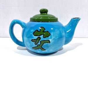 May include: A bright blue ceramic teapot with a glossy finish, featuring a dark green lid and a matching green hand-painted Japanese kanji character on the front. The teapot has a curved handle, a short spout, and a rounded body shape.