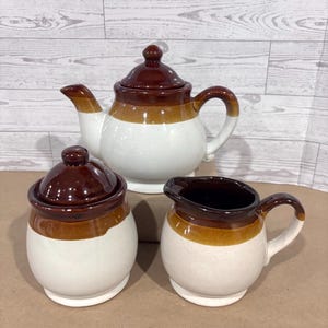 May include: A ceramic tea set consisting of a teapot with a lid, a sugar bowl with a lid, and a creamer pitcher. Each piece features a tri-color design with a dark brown top, a medium brown middle band, and a speckled off-white base.