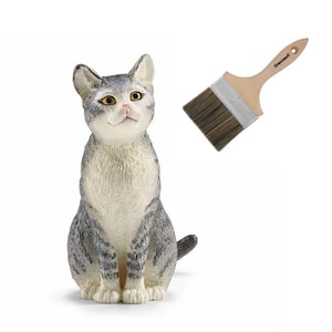Op de afbeelding: Een grijs en wit gestreept kattenbeeldje met amberkleurige ogen en een roze neus, geplaatst naast een verfkwast met een houten handvat, donkere haren en de merknaam Geninart op het handvat, alles tegen een effen witte achtergrond.