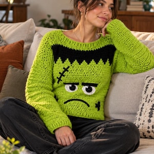 May include: A lime green long-sleeve crochet sweater featuring a black jagged neckline and a character face with white eyes, black pupils, a stitched scar, and a frowning mouth. The sweater is constructed with a textured yarn stitch pattern.