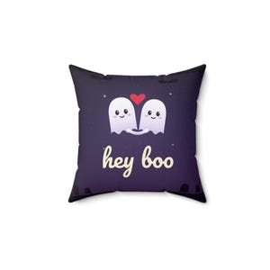 Peut inclure: Un coussin carré avec un fond violet foncé représentant deux fantômes blancs de dessin animé se tenant la main sous un cœur rouge. Le texte "hey boo" est imprimé en police cursive crème sous les fantômes, avec de petites étoiles et des chauves-souris.