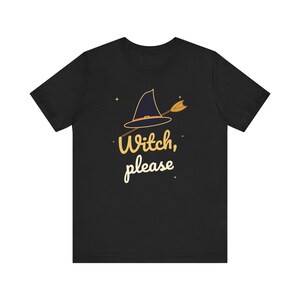 Può includere: Una t-shirt nera a maniche corte con la grafica di un cappello da strega blu scuro con una fascia arancione, trafitto da una freccia dorata. Il testo "Witch, please" è stampato in un carattere giallo chiaro sotto il cappello, con stelle.