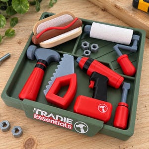 May include: A green plastic tray labelled Tradie Essentials containing a toy hammer, saw, drill, paint roller, three small nuts, and a toy hot dog. The tools are primarily red and grey, and the tray features a hammer icon logo on the front.