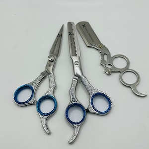 Könnte beinhalten: Ein Set aus drei silberfarbenen Friseurwerkzeugen, bestehend aus einer Haarschneideschere, einer Effilierschere mit gezahnten Klingen und einem Rasiermesser. Jedes Werkzeug hat filigrane Gravuren an den Griffen und blaue Ringeinsätze.