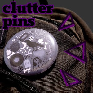 May include: A circular pin featuring a light purple background with a collage of white and black illustrations including a raven, a spider web, a skull, a cassette tape, a vinyl record, headphones, a tombstone with RIP text, and various small symbols.