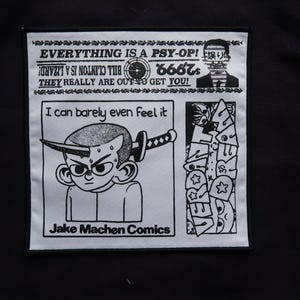 Könnte beinhalten: Ein schwarz-weißer Aufnäher mit dem Text "EVERYTHING IS A PSY-OP!" und "BILL CLINTON IS A LIZARD!" sowie einer Cartoon-Illustration einer Figur mit einem Schwert. Der Text "I can barely even feel it" ist ebenfalls vorhanden. Der Aufnäher ist mit "Jake Machen Comics" beschriftet.