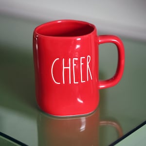 May include: A red ceramic mug with a white "CHEER" inscription. The mug is sitting on a green glass surface.