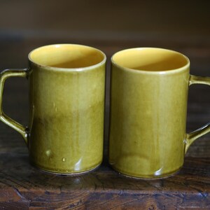 May include: Two ceramic mugs with a cylindrical shape and a handle. The exterior is a mottled olive green, while the interior is a bright yellow. The mugs are placed side by side on a dark wooden surface.