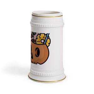 May include: A white ceramic beer stein featuring a gold rim at the top and base, with a textured beaded band near the top and bottom. The side displays a graphic of a brown skull-shaped cocktail mug with a pineapple slice, straw, and umbrella.