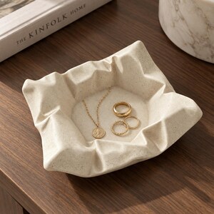 Pode incluir: Um prato quadrado, de cor branco-sujo e salpicado, com um design texturizado de bordas dobradas. Dentro do prato, repousa um colar dourado com um pingente circular e dois anéis dourados, um liso e outro com textura torcida.