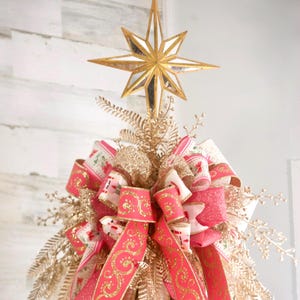 May include: A Christmas tree topper featuring a gold star and decorative bows. The star is multi-faceted and gold. The bows are pink and white with gold accents. The topper also includes gold-colored sprigs and decorative elements.