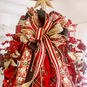 May include: A decorative Christmas tree topper featuring a gold star, layered ribbons in red plaid, solid black, and nutcracker-patterned fabric. The arrangement includes clusters of red berries, dark berries, and gold leaves, creating a full display.