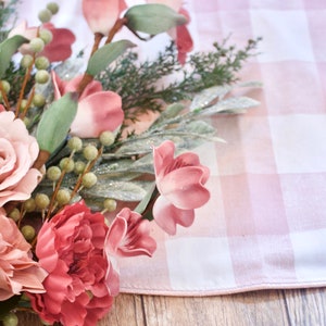 May include: A close-up of a floral arrangement featuring pink and mauve roses, peonies, and other flowers. The arrangement is set against a pink and white checkered fabric and wooden surface.