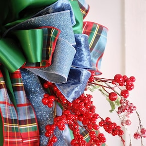 May include: A decorative bow featuring layers of solid green satin, blue glittered fabric, and a red, green, and blue plaid ribbon. The arrangement is accented with clusters of bright red berries and some frosted, snow-dusted berry branches.