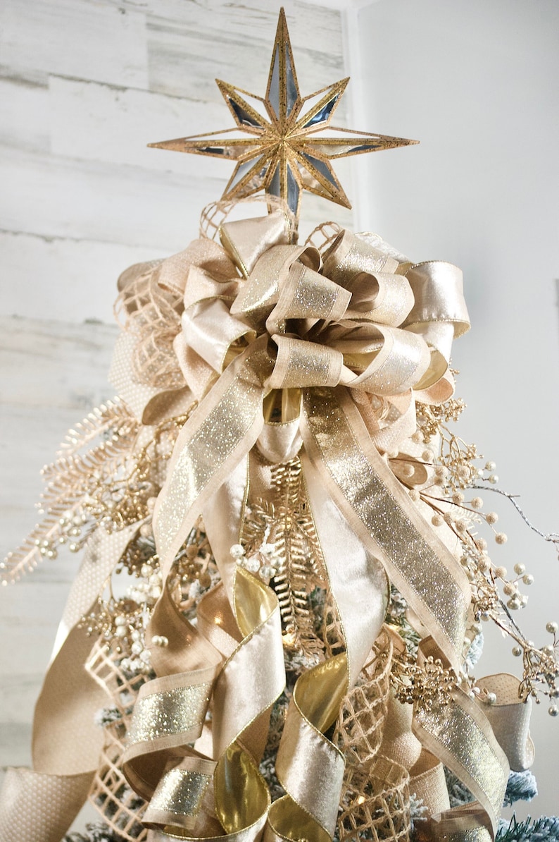 May include: A gold star tree topper with a large beige bow with gold glitter ribbon. The bow is tied around a Christmas tree with beige and gold ribbon and other decorations.