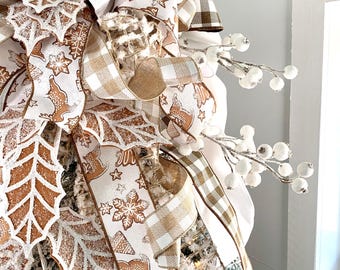 May include: A festive Christmas tree bow featuring a mix of ribbons in white, brown, and tan. The ribbons are adorned with gingerbread men, snowflakes, and star patterns. White berry sprigs and frosted leaf accents add to the holiday theme.