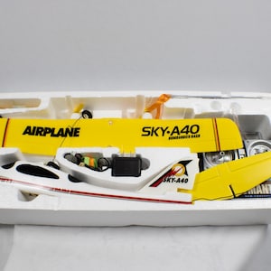 May include: A yellow and white remote-controlled aeroplane kit in a white polystyrene tray. The wings feature the text AIRPLANE and SKY-A40 BOMBARDIER DASH. The tail section includes the SKY-A40 logo. A remote control unit is included in the packaging.