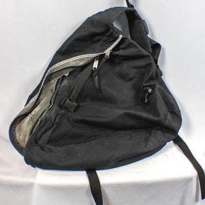 May include: A black sling backpack featuring a large triangular main compartment with a zipper closure, a black mesh front pocket, and an adjustable strap. The interior lining is a light gray fabric, and the bag includes black plastic buckles.