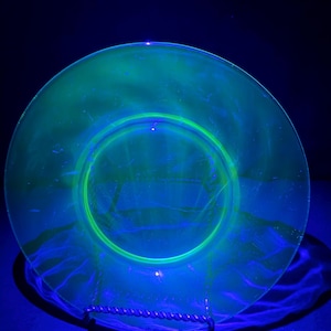 May include: A round glass plate that glows with a vibrant neon green color under ultraviolet light. The plate features a smooth, translucent surface with a circular center design and a slightly raised rim, displaying a distinct fluorescent effect.