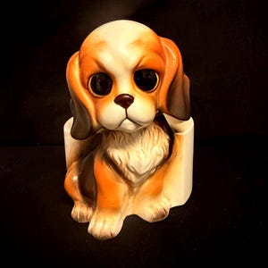 May include: A ceramic figurine of a puppy with large black eyes, long brown ears, and a white and tan coat. The puppy is sitting in front of a white cylindrical holder, featuring a smooth finish and detailed painted features on its face and chest.