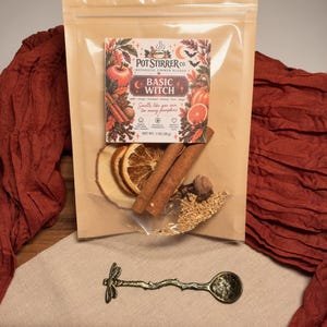 May include: A clear plastic pouch of Pot Stirrer Co. Basic Witch botanical simmer blend containing dried orange slices, cinnamon sticks, and spices, with a net weight of 1 oz. Below is a small, antique-style metal spoon with a dragonfly handle.