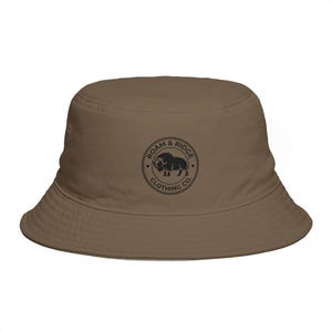 Könnte beinhalten: Ein einfarbiger brauner Fischerhut mit einem kreisförmigen schwarzen Logo auf der Vorderseite. Das Logo zeigt die Silhouette eines Moschusochsen, umgeben vom Text Roam & Ridge Clothing Co. Der Hut hat eine abfallende Krempe.