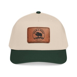 Könnte beinhalten: Eine beige Baseballkappe mit dunkelgrünem Schirm und einem dunkelgrünen Knopf oben. Ein rechteckiger brauner Lederaufnäher ist vorne mittig angebracht und zeigt ein schwarzes kreisförmiges Logo mit einem Moschusochsen, ROAM & RIDGE und EST. 2025.