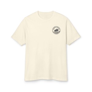 May include: A cream-colored short-sleeve t-shirt featuring a circular black and white graphic on the left chest. The graphic depicts a bison standing on a ridge with the text Roam & Ridge and Mountain Clothing Co. printed inside the circle.