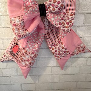 May include: A large fabric bow featuring a patchwork design of pink, white, and orange patterns including ghosts, jack-o'-lanterns, and stripes. A small patch on the lower left displays a pink ghost graphic with the text SWEET BOO.