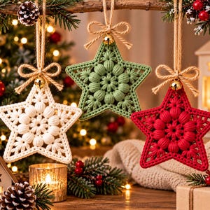 May include: Three star-shaped ornaments made of crocheted yarn in white, sage green, and red. Each star features a central puff stitch design, a small gold-colored bell, and a jute twine bow for hanging. The ornaments are suspended from a branch.