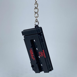 May include: A black keychain shaped like a Honda DOHC VTEC engine valve cover. The rectangular piece features the red Honda logo and the red text DOHC VTEC printed on the surface. It is attached to a silver metal split ring by a short chain.
