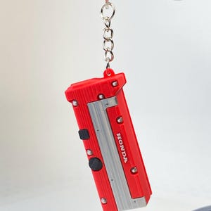 May include: A red and silver keychain shaped like a Honda engine valve cover. The rectangular red component features a central silver panel with the word HONDA printed in white, accented by small silver circular details and two black square buttons.