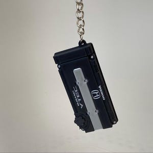 May include: A black and silver keychain shaped like a Honda DOHC VTEC engine valve cover. The rectangular piece features the Honda logo, the text DOHC VTEC, and a small black circular cap detail, all suspended from a silver metal chain link.