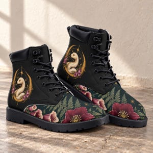 May include: A pair of black lace-up boots featuring a graphic print of a white snake coiled within a golden crescent moon, surrounded by dark green fern leaves and deep red flowers. The boots have black laces, metal eyelets, and a black sole.