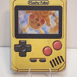 May include: A yellow handheld gaming console replica featuring the text Flashy Fabs at the top. The screen displays a holographic image of a yellow creature with red cheeks. It includes a black directional pad, two red buttons, and speaker slots.