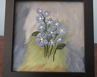 May include: A framed painting of blue forget-me-not flowers with yellow centers and green leaves. The artwork is set against a backdrop of blended gray, blue, and green hues. The painting is enclosed in a black square frame, suitable for wall decor.