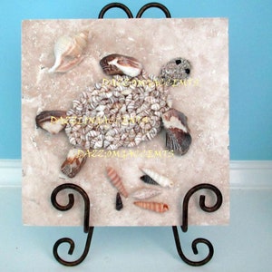May include: A decorative tile featuring a sea turtle design made from various seashells. The turtle is composed of small, white and tan shells, with larger shells forming the flippers and head. The tile is displayed on a wrought iron stand.