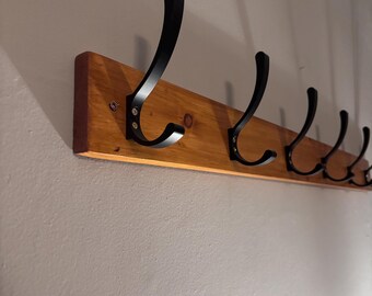 May include: A wall-mounted coat rack featuring a rectangular natural wood board with a visible grain pattern. Five matte black metal hooks are evenly spaced and securely attached to the wooden base with visible silver-toned screws.