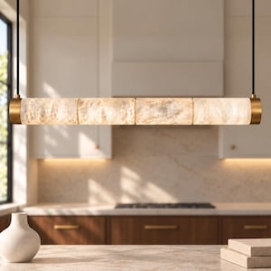 May include: A linear pendant light fixture featuring four cylindrical, textured, translucent stone-like segments illuminated from within. The light is capped with brushed brass metal ends and suspended by two thin black cords from the ceiling.