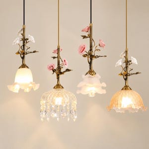 May include: Four hanging pendant lights with brass-toned stems adorned with sculpted white and pink roses. The fixtures feature varied glass shades, including smooth, frosted, and crystal-draped designs, each suspended by a thin black cord.