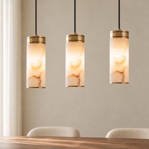 May include: Three cylindrical pendant lights hanging from black cords. Each fixture features a brushed brass metal top and base with a translucent, marbled glass shade that emits a warm, soft glow with beige and cream veining patterns.