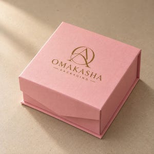 Könnte beinhalten: Eine quadratische, hellrosa Geschenkbox mit Klappdeckel. Auf der Oberseite der Box befindet sich ein goldfarbenes Logo, das aus einem stilisierten Buchstaben A in einem Kreis besteht, mit dem Text OMAKASHA PACKAGING in goldener Schrift darunter.
