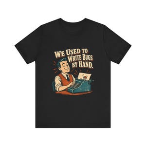 May include: A black t-shirt featuring a graphic illustration of a person typing on a blue typewriter. The text above reads WE USED TO WRITE BUGS BY HAND. The illustration includes a small red bug on the paper and uses a vintage color palette.