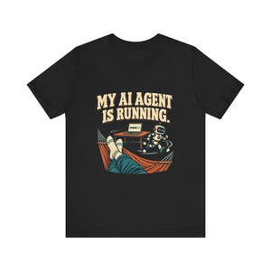 May include: A black t-shirt featuring a graphic print with the text MY AI AGENT IS RUNNING. The illustration shows a person relaxing in a hammock with feet visible, while a small robot runs near a desk with a laptop displaying a loading bar.