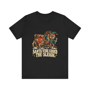 May include: A black t-shirt featuring a graphic of a robot elf holding a wrench next to a red sleigh filled with gifts. The design includes string lights and the text SANTA VIBE CODED THE SLEIGH in a retro font style at the bottom of the illustration.