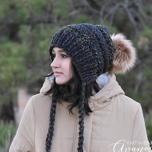 May include: A dark blue and black knit hat with a pom pom and long ear flaps. The hat is made with a chunky yarn and has a textured knit pattern. The hat is worn by a person with long dark hair and a tan jacket. The text "Knitwear Avaneska" is visible on the jacket.